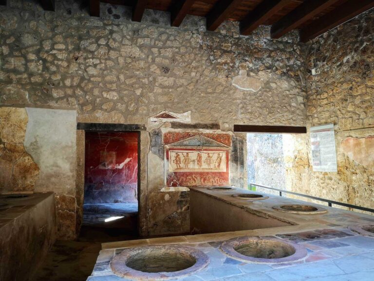 The Thermopolium of Via dell’Abbondanza in Pompeii with a bar in the foreground with sunken jars in it for food and a fresco on the back wall